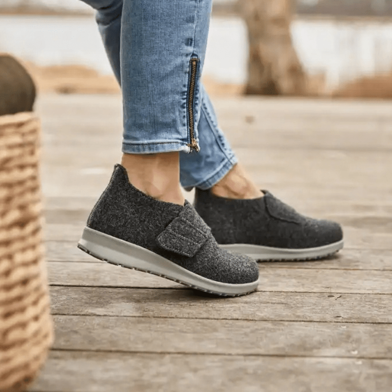 Dame iført antrasittgrå New Feet hjemmesko på et tregulv, sett fra siden med fokus på føttene