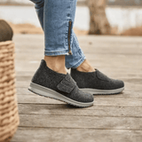 Dame iført antrasittgrå New Feet hjemmesko på et tregulv, sett fra siden med fokus på føttene