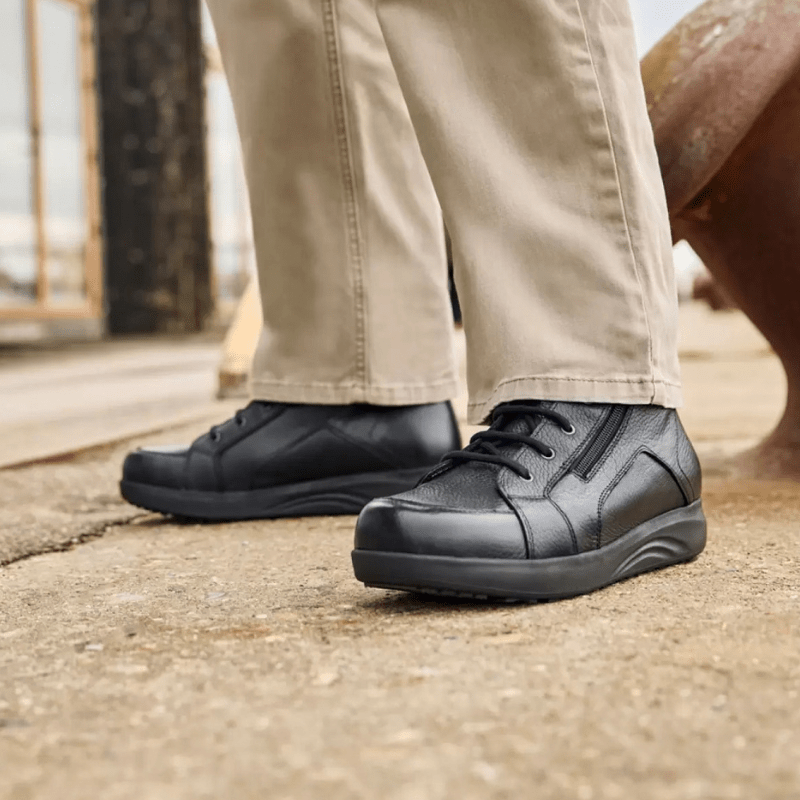 En mann iført beige bukser og svarte New Feet herrestøvler står på et grått underlag